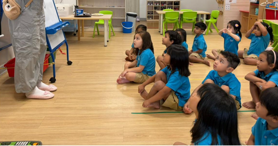 Children in blue uniforms sit on a classroom floor, watching a teacher beside a whiteboard.